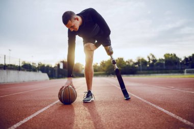 Full length of sporty caucasian handicapped man in sportswear and artificial leg dribble the ball while standing on racetrack. clipart