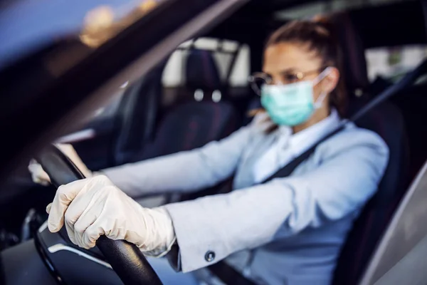 Businesswoman with mask and gloves on driving her car. Hands are on steering wheel. Selective focus on hand. Corona virus protection concept.