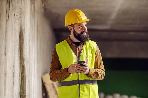 年轻英俊的胡子工人站在水泥墙前 用智能手机 — 图库照片