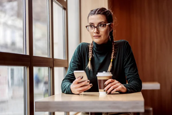 Jovem Mulher Pensativa Sentada Cafetaria Segurando Copo Telefone Inteligente Enquanto — Fotografia de Stock