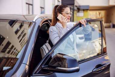 Young attractive businesswoman dressed smart casual exiting her car and having phone conversation. clipart