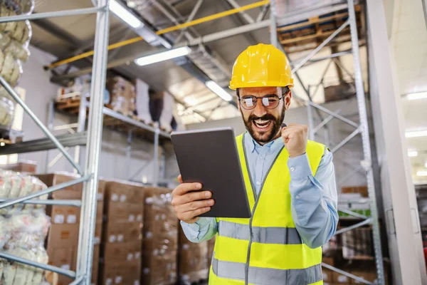Young smiling supervisor in vest and protective helmet standing in warehouse and using tablet for work. Salary is going well so he is excited.