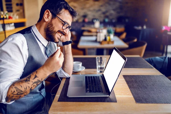 Side view of handsome Caucasian bearded businessman with tattoo and eyeglasses in suit sitting in cafe, putting earphones in ears and preparing to have conference call.