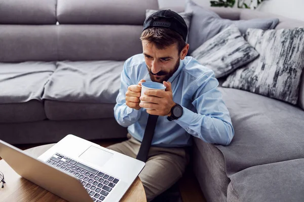 若いビジネスマンは床の上の家に座ってコーヒーを飲みながらノートパソコンを見て — ストック写真