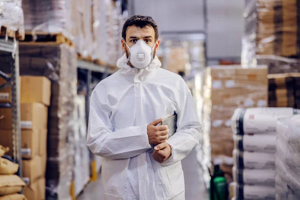 Food factory worker in protective sterile suit and mask staining in warehouse and holding tablet under armpit.