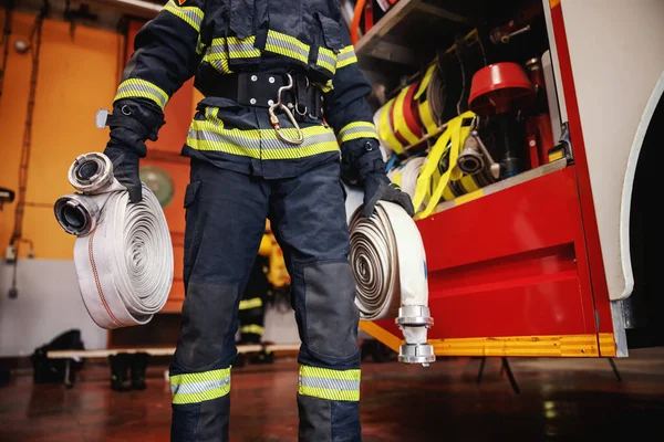 Firefighter in protective uniform with helmet on head checking on hoses before intervention while standing in fire station.