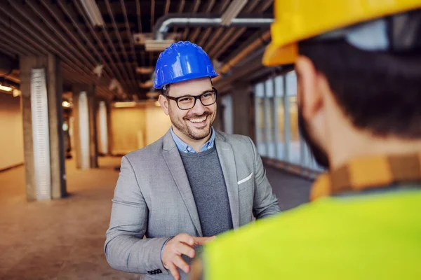 建設プロセスの建物に立っている間 請負業者と話を頭の上にヘルメットに合わせて笑顔建築家 — ストック写真