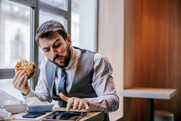 Hungry man in suit sitting in fast food restaurant on lunch break, eating cheese burger and reading news on tablet.