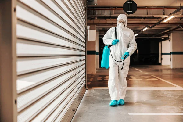 完全な長さの男で保護滅菌均一運搬噴霧器で消毒剤とガレージを歩く — ストック写真
