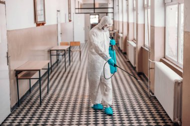 Worker in sterile white uniform, with mask and glasses holding sprayer with disinfectant and spraying around hallway in school. Prevention of spreading corona virus. clipart