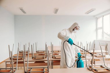 Man in sterile uniform, with gloves and mask holding sprayer and spraying with disinfectant desks and chairs in classroom. clipart