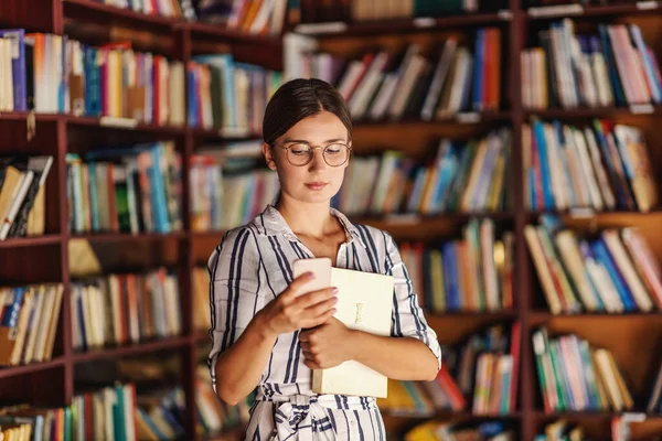 Młoda Atrakcyjna Studentka Stojąca Bibliotece Trzymająca Książkę Korzystająca Smartfona — Zdjęcie stockowe