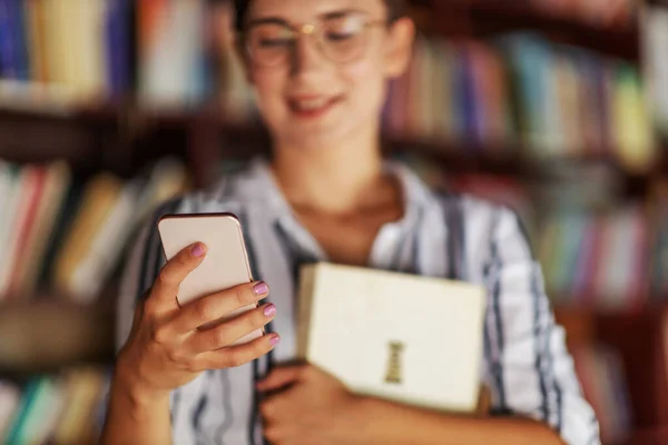 Zbliżenie Młodej Atrakcyjnej Studentki Stojącej Bibliotece Trzymającej Książkę Korzystającej Smartfona — Zdjęcie stockowe