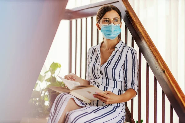 Young dedicated intelligent college girl with face mask on sitting on stairs and reading a book during corona virus pandemic.