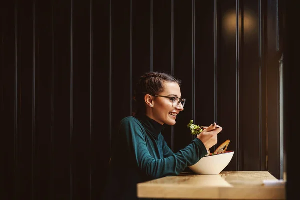 ウィンドウの近くのレストランに座って 昼食のためのサラダを持つ健康的な習慣を持つ若い女性の側面図 — ストック写真