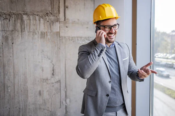 スーツを着た笑顔の建築家と頭の上にヘルメット窓の横に建設プロセスの建物に立って スマートフォンで話して — ストック写真