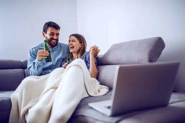かわいい陽気な白人のカップルは リビングルームでソファに座ってブランケットで覆われて ビールを飲み ラップトップ上で映画を見ます — ストック写真