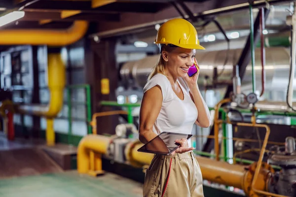 Middle aged smiling female supervisor in working uniform with helmet on head holding tablet and having phone conversation while standing in heating plant.