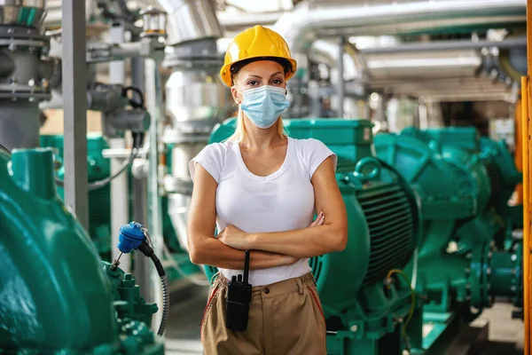 Hardworking successful independent female worker with protective helmet in working uniform and with face mask on standing in heating plant with arms crossed during corona virus outbreak.