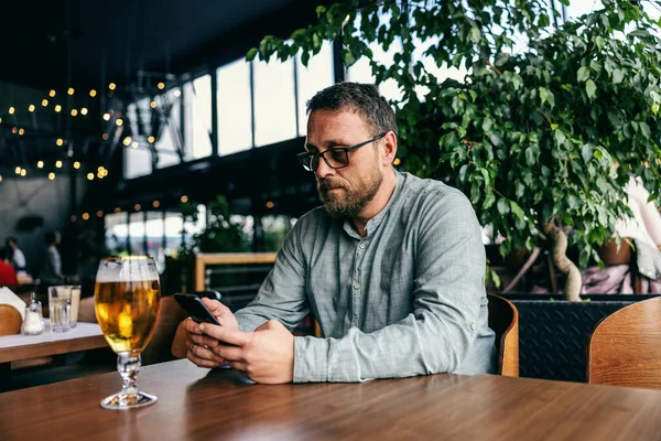 Middle aged bearded man sitting in a bar and using smart phone for hanging on social media sites. In front of him on a table is a cold fresh beer.