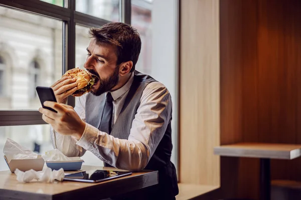 Éhes Üzletember Ebédszüneten Gyorsétteremben Sajtburgert Eszik Okostelefont Használ — Stock Fotó