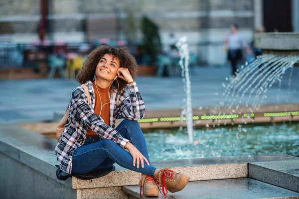 年轻迷人的混血女子坐在喷泉上欣赏音乐 — 图库照片