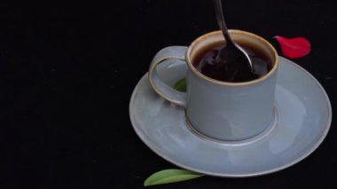 4k video Yakın karıştırın veya yeşil yaprak ve kırmızı gül yaprağı ile siyah arka plan üzerinde bir kaşık ile mavi bir kahve fincanı karıştırma ve daha sonra aşağı döşeme