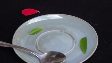 4k video Yakın karıştırın veya yeşil yaprak ve kırmızı gül yaprağı ile siyah arka plan üzerinde bir kaşık ile mavi bir kahve fincanı karıştırma ve daha sonra aşağı döşeme