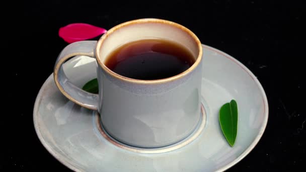 Vidéo 4k Prendre la cuillère et remuer ou remuer une tasse de café bleu avec une cuillère sur fond noir avec feuille verte et pétale de rose rouge 