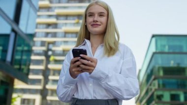 Beyaz tişörtlü, akıllı telefon tutan ve gülümseyen genç profesyonel bir kadın modern kentsel iş bölgesinde açık havada mesajlaşırken....