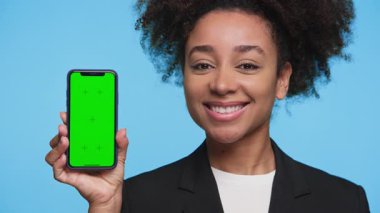 Neşeli genç profesyonel kadın elinde yeşil ekranlı bir akıllı telefon tutuyor, özelleştirme ya da uygulama sunumuna hazır, mavi arka planda izole edilmiş..