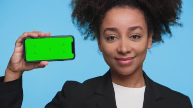 Elinde yeşil ekranlı akıllı bir telefonla gülümseyen profesyonel bir kadın, kafasını hafifçe sallıyor, sanki bir şeyi onaylıyor ya da onaylıyormuş gibi, mavi arka planda izole edilmiş..