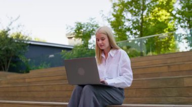 Odaklanmış profesyonel bir kadın yeşil bir alanda ahşap basamaklara oturur ve rahat bir uzaktan kumandada dizüstü bilgisayarı üzerinde çalışır..
