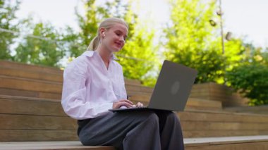 Rahat bir kadın, doğal ortamında, tahta merdivenlerde oturur. İnterneti kablosuz kulaklıklarıyla gezer..