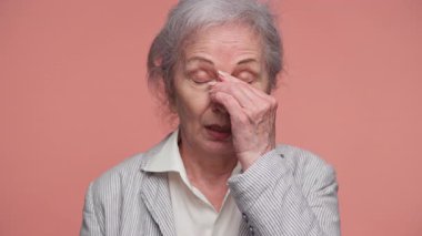 Çizgili ceketli yaşlı kadın alnını tutuyor ve gözlerini kapatıyor, baş ağrısını ve rahatsızlığını gösteriyor, pembe arka planda soyutlanmış..