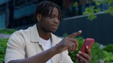 Dışarıda akıllı telefonunu kullanan genç bir adamın görüntüsü. Sıradan bir ortamda, odaklanmış bir ifadeyle ekranda kayıyor..