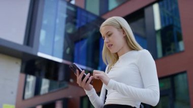 Modern bir binanın önünde duran genç bir kadın, akıllı telefonuna bakıp yüzünü kapatırken gözle görülür şekilde üzgün ya da stresli..