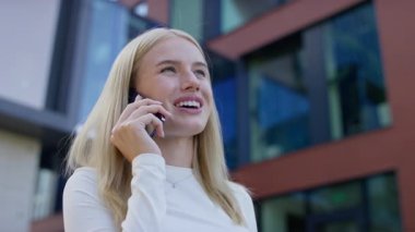 Gülümseyen genç bir kadın güneşli bir günde modern bir şehir ortamında dışarıda beklerken keyifli bir telefon konuşması yapıyor..
