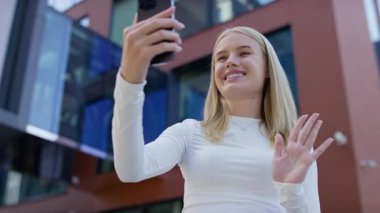 Gülümseyen genç bir kadın akıllı telefon tutuyor ve video görüşmesi sırasında el sallıyor, modern bir şehir ortamında açık havada duruyor..