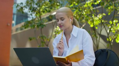Kulaklık takan ve elinde bir not defteri olan genç bir kadın açık hava ortamında çevrimiçi bir derste dizüstü bilgisayarını dikkatle izliyor..