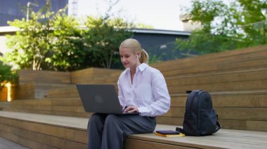 Kulaklık takan sıradan bir genç kadın yeşil ve güneş ışığıyla çevrili şehir parkındaki ahşap merdivenlerde otururken dizüstü bilgisayar kullanıyor..