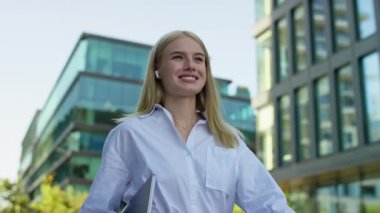 Modern bir şehirde elinde dizüstü bilgisayarla dolaşan, kulaklık takan ve beyaz gömlek giyen gülümseyen genç bir profesyonel..