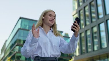 Kulaklık takan ve video görüşmesinde el sallayan gülümseyen genç bir kadın, yüksek binaları olan modern bir şehirde yürüyor..