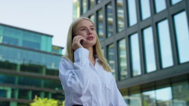 Beyaz tişörtlü, kendinden emin genç bir kadın modern bir şehirde cam binalarla yürürken akıllı telefonuyla konuşuyor..