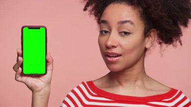 Kırmızı çizgili gömlekli genç bir kadın, yeşil ekranlı, pembe arka planda izole edilmiş bir telefonu gösterirken başını hafifçe sallıyor.