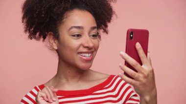 Kırmızı çizgili tişörtlü mutlu genç bir kadın video görüşmesi sırasında konuşuyor, telefonunu tutuyor ve pembe bir arka plana gülümsüyor..