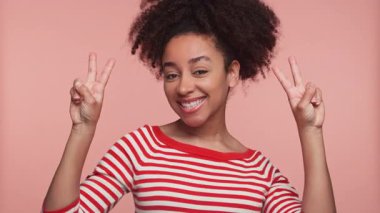 Kırmızı çizgili tişörtlü neşeli genç kadın kameraya gülümserken iki elini de pembe arka planda izole edilmiş barış işareti hareketleriyle kaldırıyor..