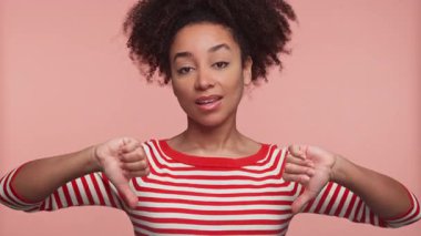 Kırmızı çizgili gömlekli genç bir kadın, pembe arka planda izole edilmiş bir şekilde stüdyoda onaylamadığını ya da olumsuz tepkiler verdiğini belirterek iki kez baş parmak hareketi yapıyor..