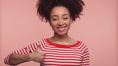 Kırmızı çizgili tişörtlü mutlu genç kadın pembe stüdyo geçmişine başparmak işareti yaparken kendine güvenerek gülümsüyor..