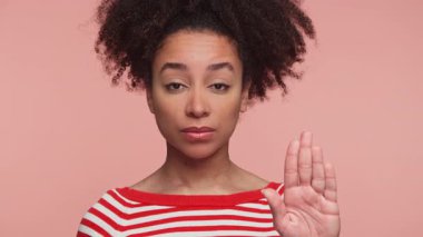 Kırmızı çizgili tişörtlü genç bir kadın elini ileri doğru tutarak pembe bir arka plana karşı ciddiyetini ifade ediyor..